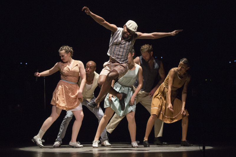 Byron Tittle, in shorts, a newsboy cap and vest jumps high into the air. The ensemble in mismatched dresses and vests stand behind in a clump with smirks on their faces.